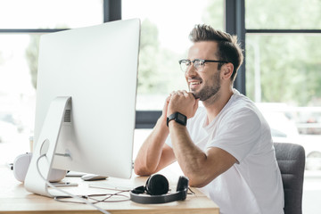 smiling young programmer in eyeglasses using desktop computer at workplace