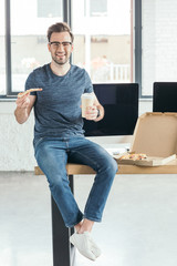 handsome smiling young programmer in eyeglasses holding paper cup and eating pizza at workplace
