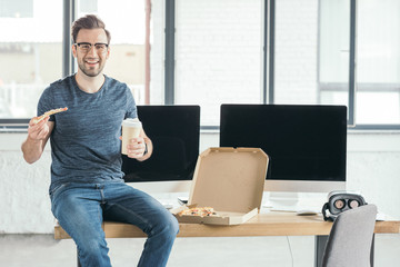 smiling young programmer in eyeglasses holding paper cup and eating pizza at workplace