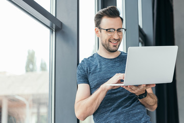 smiling young man in eyeglasses using laptop near window