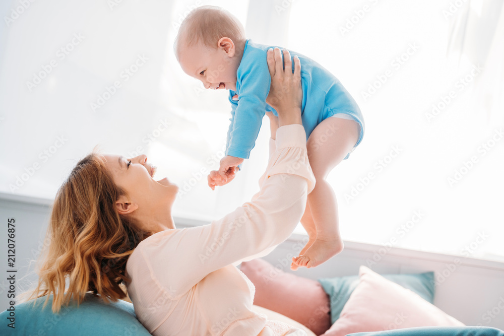 side view of happy mother raising laughing little child while sitting on bed at home - obrazy, fototapety, plakaty 
