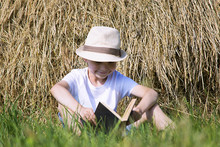 Cute Little Boy Is Reading A Book In A Straw Stack On The Grass In Summer