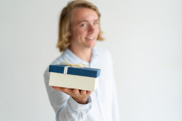 Wall Mural - Smiling handsome guy offering gift. Blonde Caucasian man out of focus holding present box in focus. Holiday, festival, special day concept