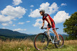 Athletic sportsman cyclist in sportswear and helmet riding a bicycle in high grass. Active male biker enjoying beautiful view of mountains on summer day. Healthy lifestyle and extreme sport concept