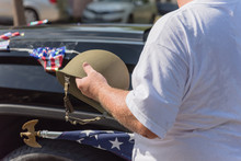 Close-up White Veteran Proudly Holding Military WWI Helmet (M1 Helmet) And US Flag During Parade. July 4th Or Veterans Day Poster Of WWII, Modern Wars. American Soldier Troop Background, Decorated Car