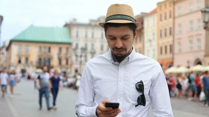 young walking stylish guy using smart phone mobile on crowded city square with blur background summer casual fashion straw hat sunglasses and shirt checking message notification young successful man