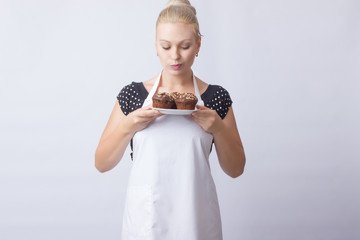 Wall Mural - A happy blond woman in a white apron shows a chocolate cupcakes on a white plate. Cook. A housewife