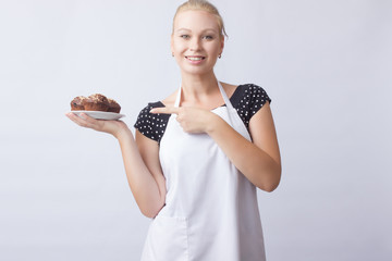 Wall Mural - A happy blond woman in a white apron shows a chocolate cupcakes on a white plate. Cook. A housewife