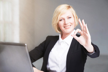 Pretty blonde businesswoman dressed in black suit shows OK sign at workplace