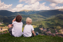 Two Boys Sitting On A Hill And Looking At The Mountains. Back View