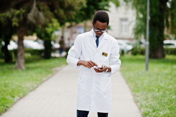 Wall Mural - Stylish african american doctor with stethoscope and lab coat, at glasses posed outdoor and using his mobile phone.