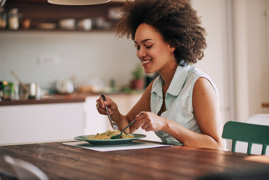 beautiful mixed race woman eating pasta for dinner while sitting at kitchen table.