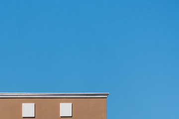 Close up in a partial geometric design of a top part of a modern building colored with a brown wall and details in white, along with a large space of a blue sky