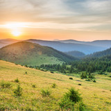 Fototapeta Na ścianę - Sunset in the mountains with forest, green grass and big shining sun on dramatic sky
