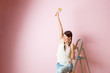 Image of cheerful woman with brush next to stepladder and roller against blank pink wall