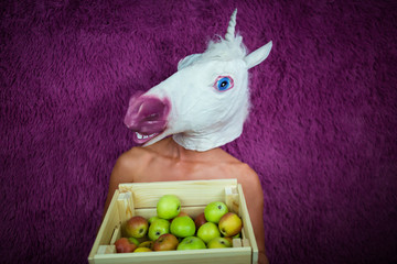 Freaky young woman in comical mask stands on the purple background and shows box of apples. Funny girl unicorn holds case with fruits in hand. Portrait of unusual lady.