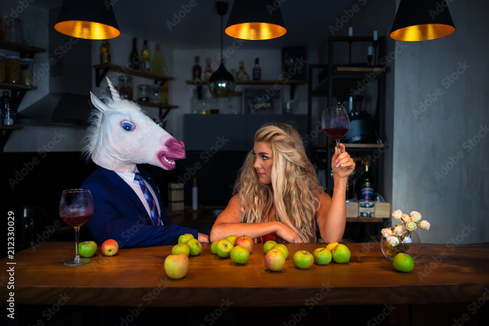 Unusual couple spend time together at the bar counter in stylish apartments with wine and food. Beautiful girl relaxing with funny boyfriend in comical mask. Unicorn in suit with young woman - obrazy, fototapety, plakaty 