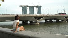 Woman Sits On A Parapet On The River Embankment Overlooking The Marina Bay Sands Hotel In Singapore
