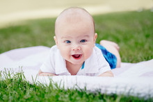 Closeup Portrait Of Adorable Funny White Caucasian Baby Boy With Blue Grey Eyes Lying On Grass In Park. Aware Cute Newborn Child On White Background Outdoors.