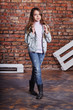 Hipster girl child wearing white cotton t-shirt, denim jacket,jeans posing against rough brick wall, minimalist urban clothing style. Full-length kid teenager model in stylish casual clothes.
