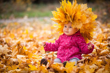 Little Kid Is Playing And Sitting In Fallen Leaves In Autumn Park. Baby Is In Big Wreath Of Leaves. Girl Is Dressed In Warm Hat, Jacket.