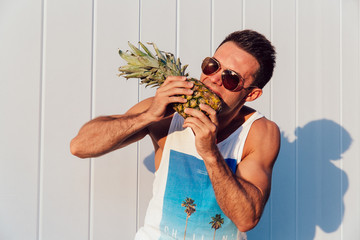 Wall Mural - Stylish muscular man wearing sunglasses trying to eat pineapple, standing outdoors. Summer time concept.