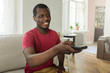 © Damir Khabirov - Horizontal photo of African American man sitting on sofa at home watching television, holding remote control in hand, pulling arm forward to switch channels with friendly relaxed smile, having rest