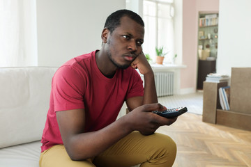 Indoor picture of young African man staying at home in front of TV set with remote control in hand, pressing fist to chin in gesture of boredom, looking indifferent towards what he sees on screen