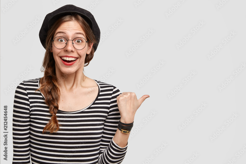 Happy Parisian female with positive smile, indicates aside at blank space, being amazed by something, isolated over white background. Cute French woman in beret and spectacles has broad smile - obrazy, fototapety, plakaty 