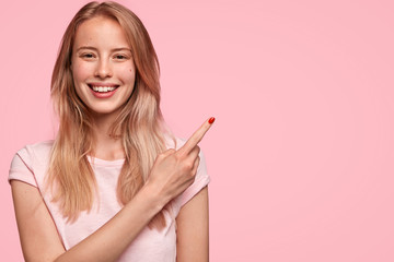 Wall Mural - Happy young cute female with cheerful positive expression, dressed in casual light pink t shirt, indicates at upper right corner, isolated over pink background, advices to go in this direction