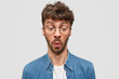 © wayhome.studio  - Indoor shot of surprised unshaven male wonders while notices something down, has displeased expression, wears glasses and shirt, stands alone against white background. Omg and reaction concept