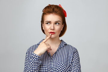 Wall Mural - Body language. Thoughtful uncertain pretty girl wearing vintage hairstyle and bright make up holding finger on her lips and looking sideways, feeling doubtful about something, posing in studio