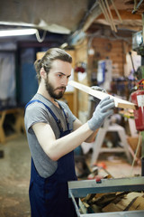 Wall Mural - Young gloved worker looking at wooden workpiece before processing it in machine