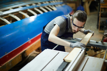 Wall Mural - Young woodworker in workwear, gloves and eyeglasses putting wooden board into machine for processing