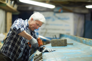 Wall Mural - Senior gloved worker with power drill leaning over large vessel and drilling one of its boards