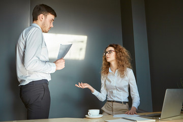 Two colleagues discussing project in office. Attractive experienced woman financial expert in formal wear and glasses sitting at her workplace, talking to her self conscious male assistant with papers