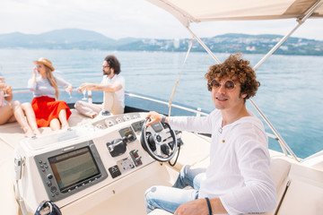 Young cheerful curly guy navigating the sailing boat in company of his friends dressed in marine style spending their weekend at sea - Concept of friendship and travel with best friends.