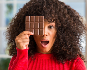 Wall Mural - African american woman eating chocolate bar at home scared in shock with a surprise face, afraid and excited with fear expression