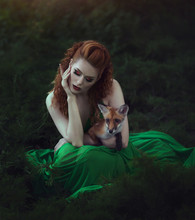 A Red-haired Girl In A Green Dress Is Sitting With A Fox In A Fairy Forest. Beautiful Young Woman With A Young Fox.