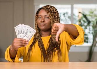 Sticker - African american woman holding dollars with angry face, negative sign showing dislike with thumbs down, rejection concept