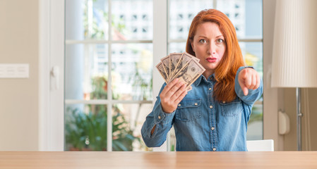 Wall Mural - Redhead woman holding dollar bank notes at home pointing with finger to the camera and to you, hand sign, positive and confident gesture from the front