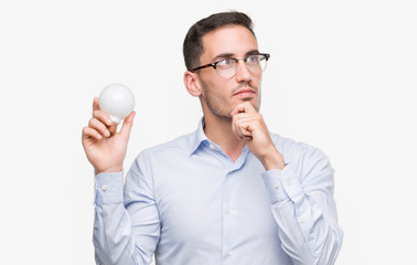 Poster - Handsome young man holding lightbulb as idea serious face thinking about question, very confused idea