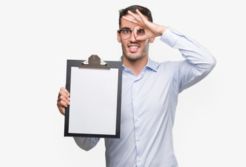 Sticker - Handsome young business man showing a clipboard with happy face smiling doing ok sign with hand on eye looking through fingers