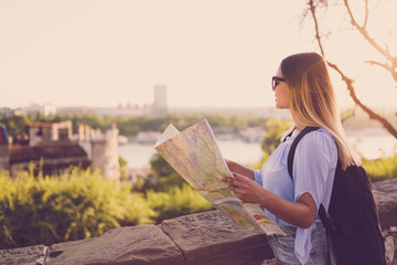 Wall Mural - Young female traveler standing in front of beautiful city view and looking at the map 