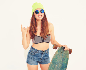 Wall Mural - Young stylish smiling girl model in casual summer swimwear clothes and yellow beanie posing with longboard desk, showing rock and roll sign. Isolated on white