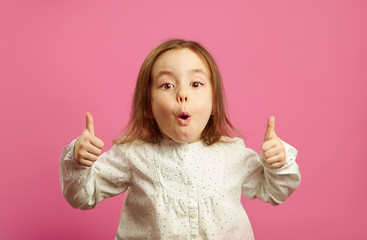 Wall Mural - Portrait of emotional little girl showing thumbs up, has surprised expression, opened her mouth, expresses astonishment and unexpected joy, standing on pink isolated background.