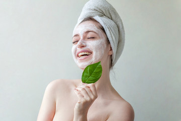 Wall Mural - Beauty portrait of a smiling brunette woman in a towel on the head with white nourishing mask or creme on face and green leaf in hand on white background isolated. Skincare cleansing eco organic