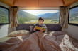 © zephyr_p - Young Asian man staying in the blanket reading magazine book in camper van with mountain scenery through the window. Road trip in summer od South Island, New Zealand.