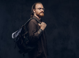 Wall Mural - Portrait of a redhead hipster student in glasses dressed in a brown shirt, holds a backpack, posing at a studio. Isolated on the dark background.