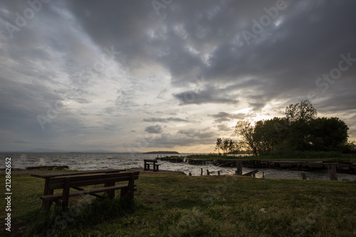 Fotografie, Obraz A wooden sitting bench on a lake shore at sunset, beneath a moody, cloudy sky